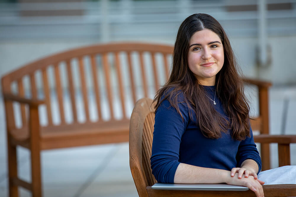 Lander College for Women student Chana Chambre sitting in a wooden chair outside on the LCW patio. She's wearing a blue shirt and has long brown hair. 