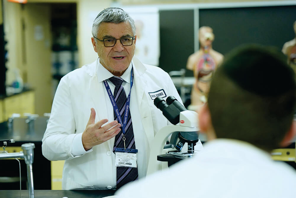 Male professor with gray hair, glasses, white lab coat and tie, gesturing with his hands, speaking to a student inside a science laboratory setting.